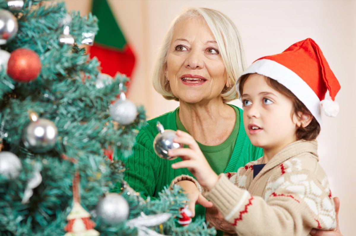 Ein kleiner Junge mit Weihnachtsmann-Mütze schmückt mit seiner Oma einen Weihnachtsbaum.